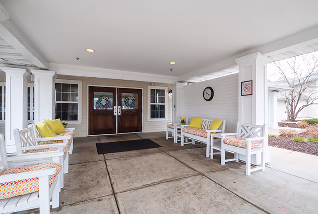 Covered entrance area of a senior living facility with white wooden chairs and benches featuring colorful cushions and yellow pillows. Double wooden doors with decorative glass panels are centered in the background. A clock and a no smoking sign are mounted on the white wall to the right. The area is clean and well-lit with recessed ceiling lights.