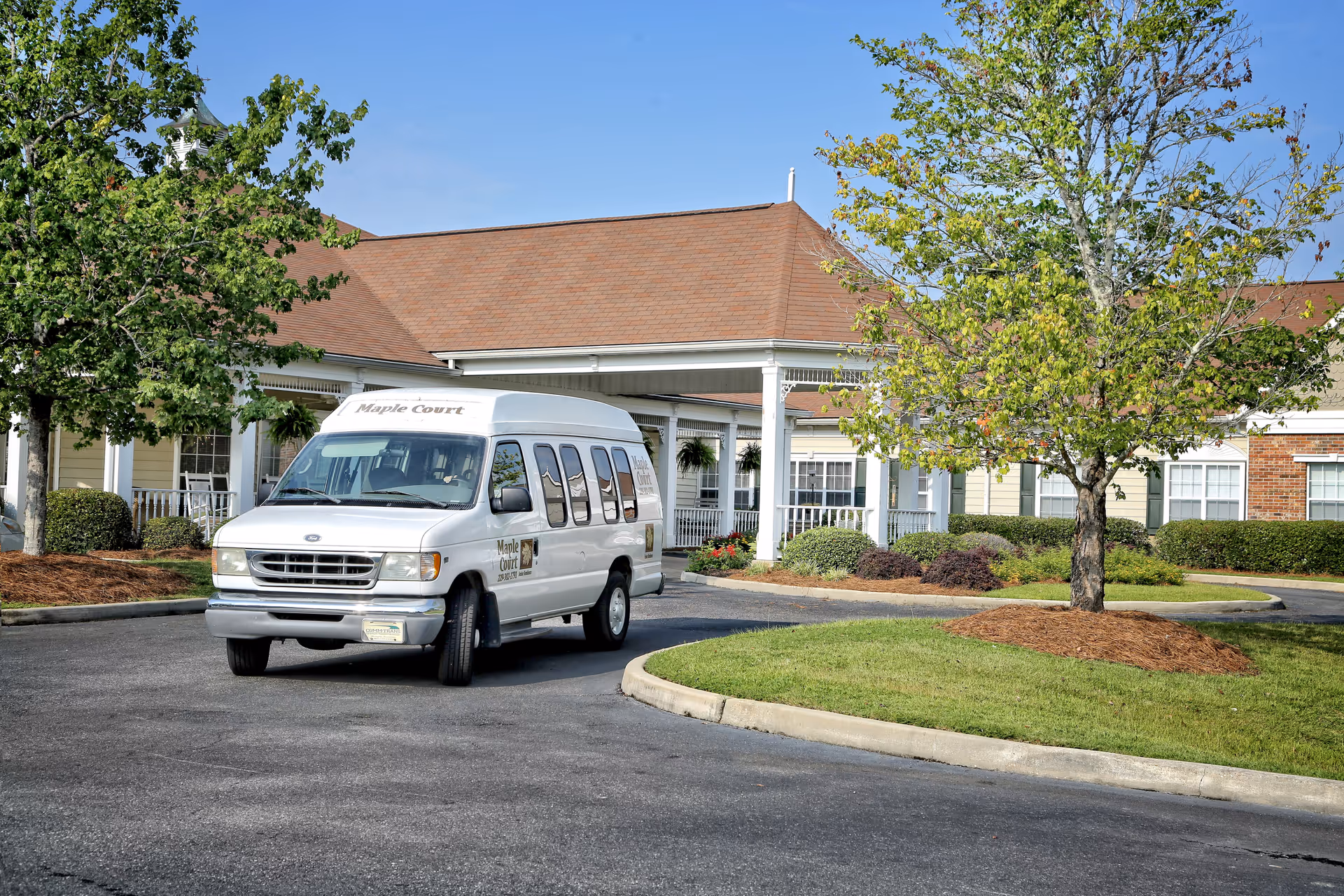 A white Maple Court senior care shuttle van parked in front of a single-story senior living building with a covered entrance and landscaped grounds.