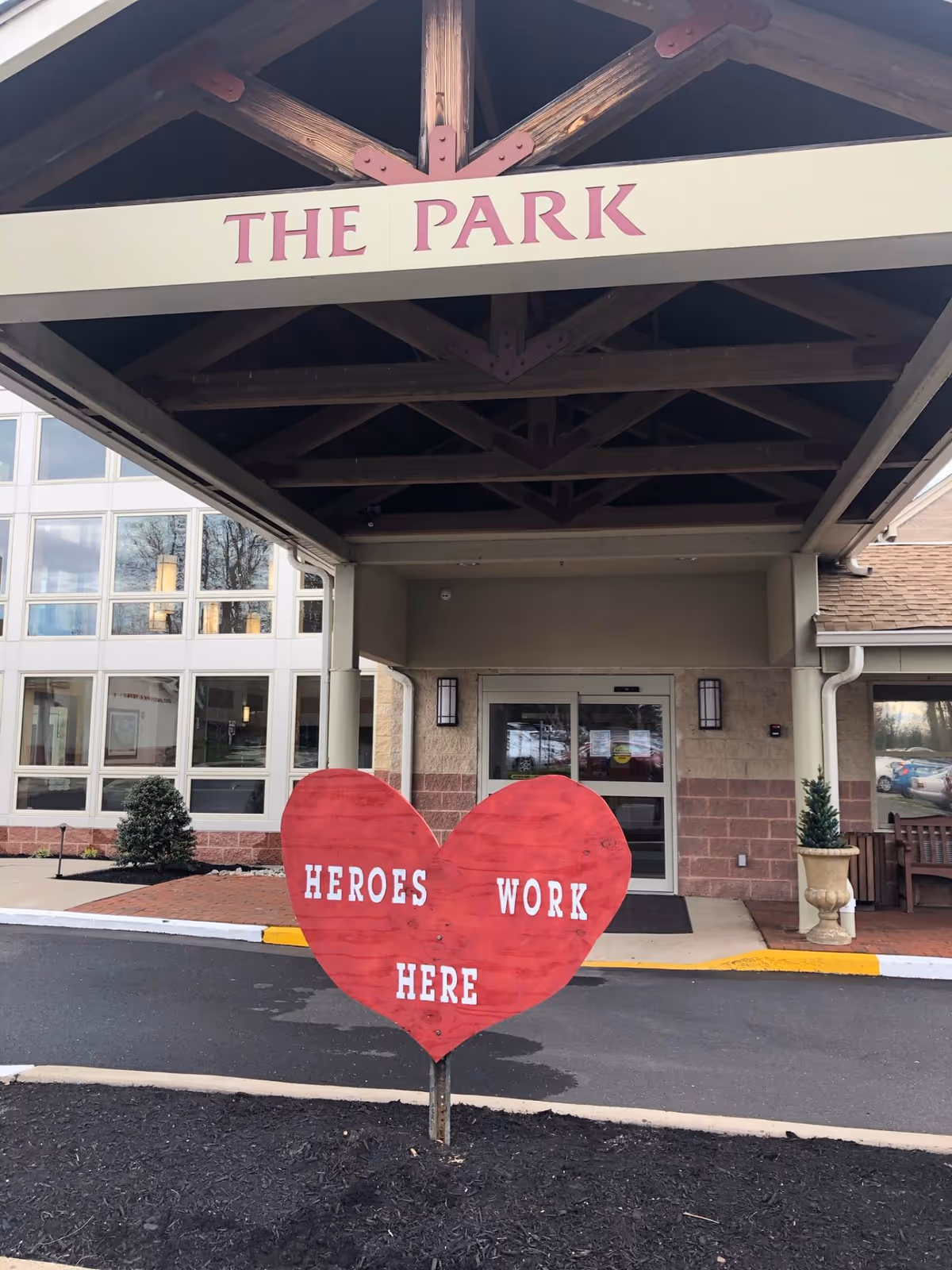 Entrance to a building with a covered driveway labeled 'THE PARK'. In front of the entrance is a large red heart-shaped sign with the words 'HEROES WORK HERE'. The building has large windows and a double glass door entrance.