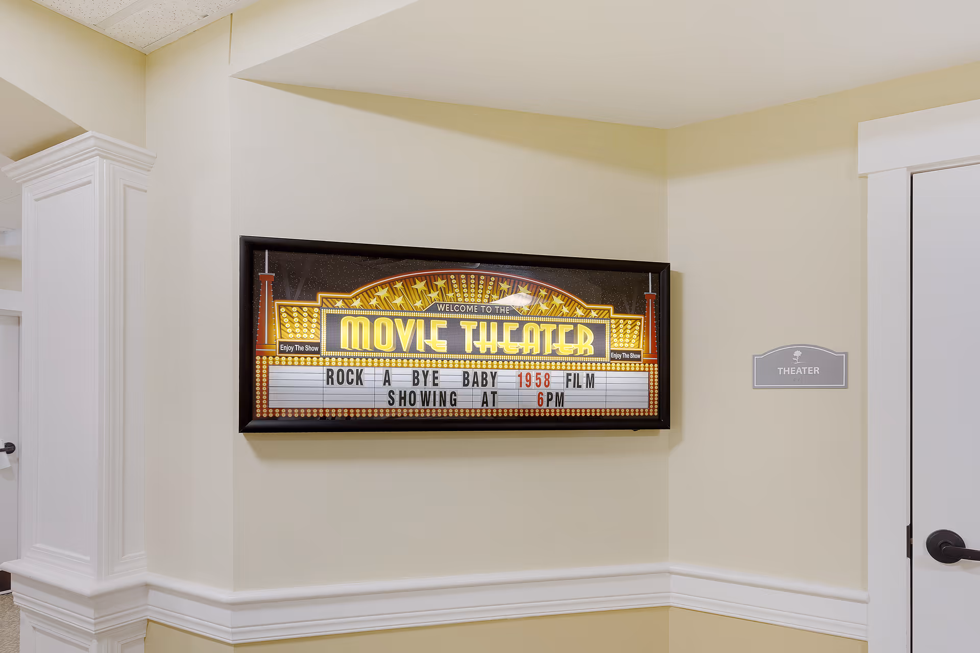 Interior wall corner with a framed illuminated sign reading 'Welcome to the Movie Theater' and a message about the film 'Rock A Bye Baby' showing at 6 PM. A small sign next to a white door reads 'Theater'.