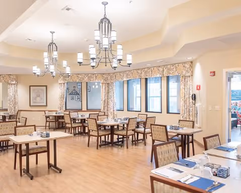 A bright dining room with multiple wooden tables and chairs arranged neatly. The room features large windows with floral curtains, modern chandeliers hanging from the ceiling, and place settings on the tables including blue placemats and napkins.