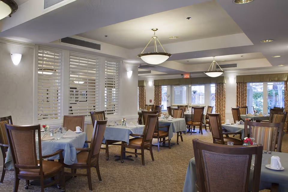 A well-lit dining room with multiple tables covered in light blue tablecloths, each set with napkins, glasses, and small flower arrangements. The room features wooden chairs with cushioned seats, large windows with curtains, and ceiling light fixtures providing warm lighting.