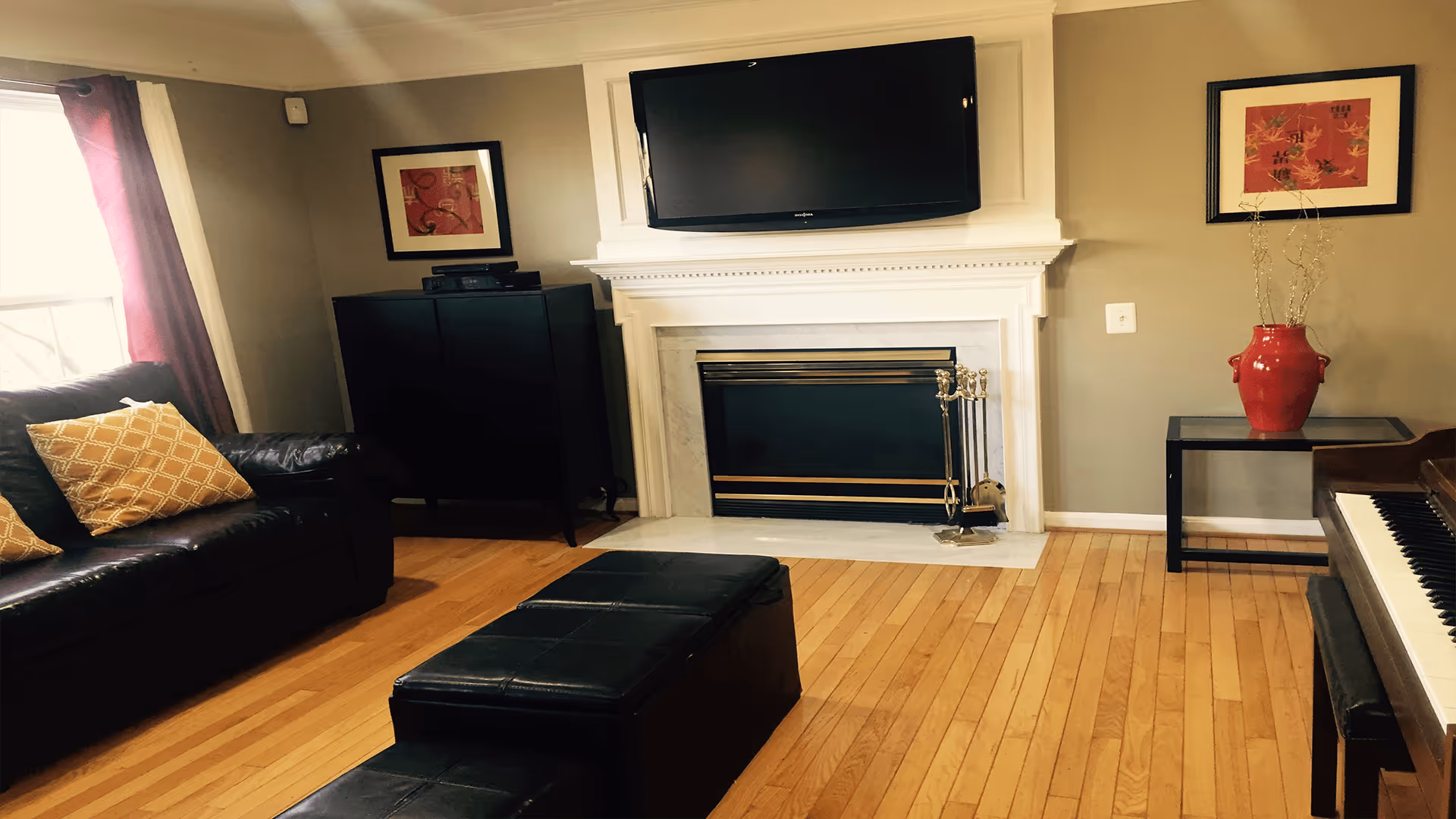 A cozy living room with hardwood floors featuring a black leather sofa with yellow patterned pillows, a black ottoman in the center, a fireplace with a mounted flat-screen TV above it, a black cabinet, a side table with a red vase, and a piano on the right side.