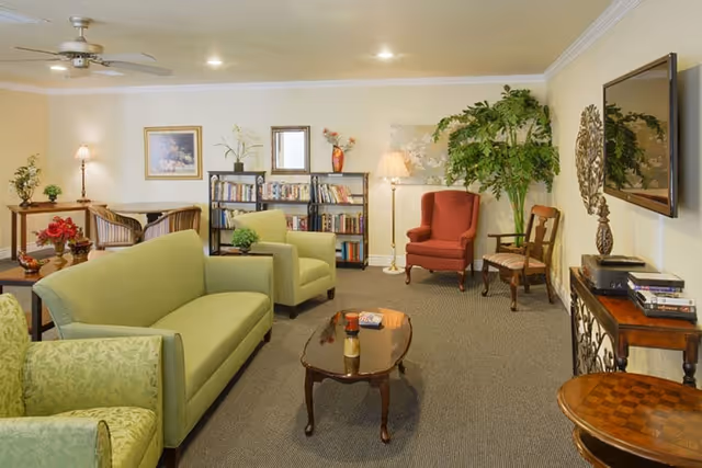 A cozy living room with light green sofas and armchairs, a red armchair, wooden chairs, a coffee table, a bookshelf filled with books, a flat-screen TV mounted on the wall, decorative plants, and warm lighting from lamps.