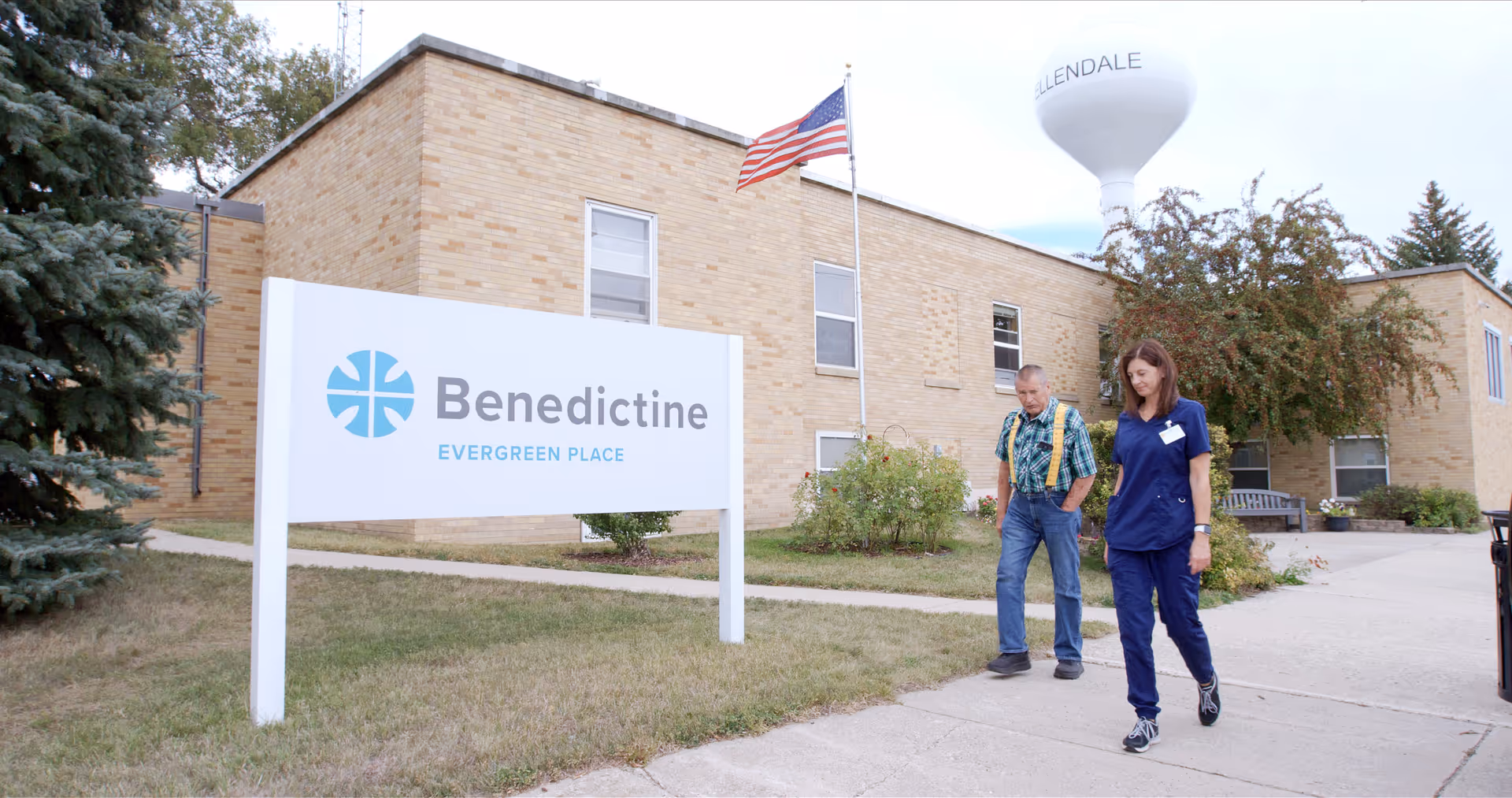 Exterior view of the Benedictine Evergreen Place sign in front of a brick building with two people walking on the sidewalk.