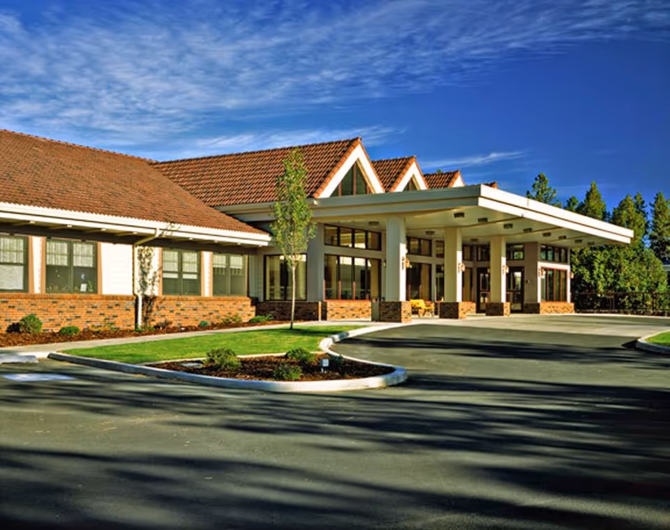Front entrance of a low-rise senior living building with a covered porte-cochère, brick façade, and landscaped driveway under a blue sky.
