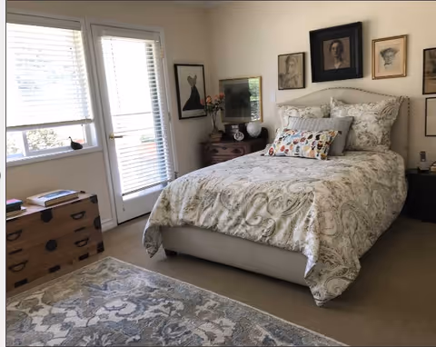 A cozy bedroom with a neatly made bed featuring patterned bedding and decorative pillows. The room has a light-colored carpet and a patterned area rug. There is a wooden chest with books on it near a window with blinds and a glass door leading outside. The walls are adorned with framed artwork above the bed and near the corner nightstand, which holds a vase with flowers and other decorative items.