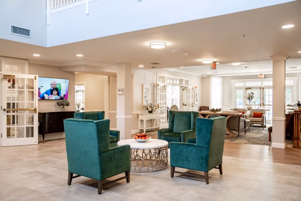 Spacious senior living lounge with four teal armchairs arranged around a round coffee table and additional seating and a TV in the background.