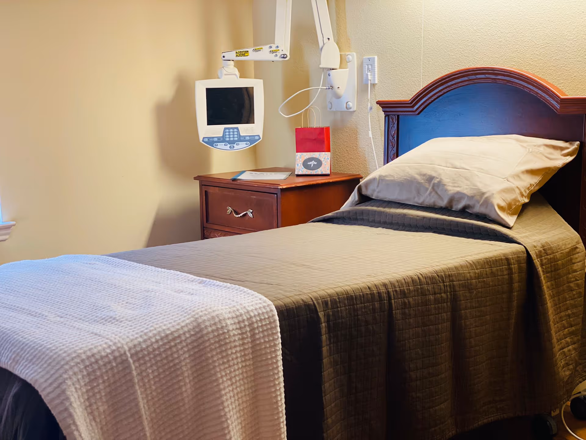 Single twin bed with a wooden headboard, pillow and blanket next to a nightstand and a wall-mounted medical monitor.