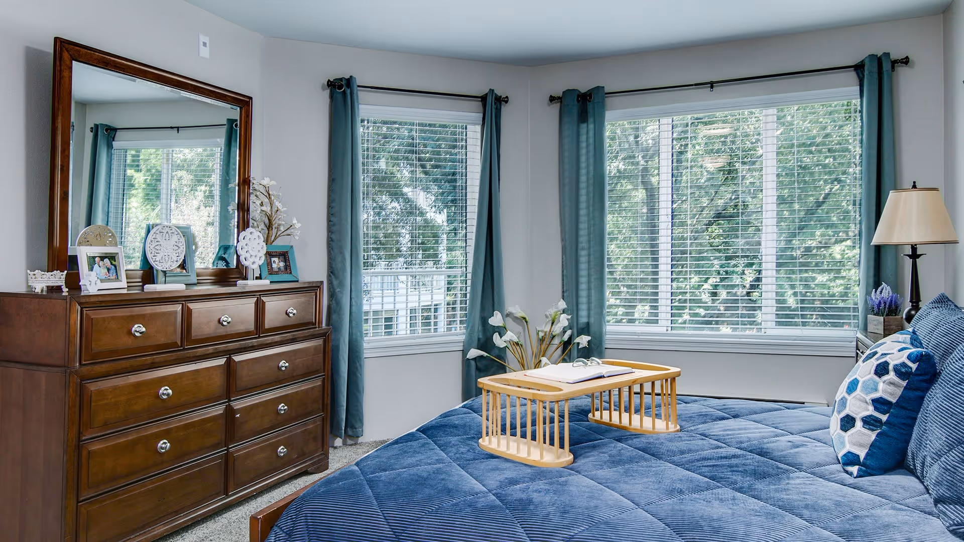 A bright bedroom with large windows covered by teal curtains, a wooden dresser with a large mirror and decorative items on top, and a bed with a blue quilted bedspread and patterned pillows. A wooden tray with an open book is placed on the bed.