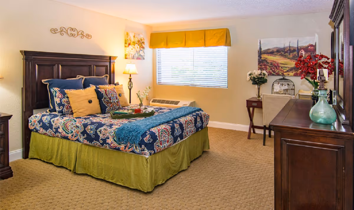 A cozy bedroom with a large bed featuring a colorful patterned bedspread and multiple pillows. The room has beige walls, a window with a yellow valance, a wooden headboard, a nightstand with a lamp, a dresser with decorative items, and a small desk with a chair. There are paintings and floral decorations adding warmth to the space.
