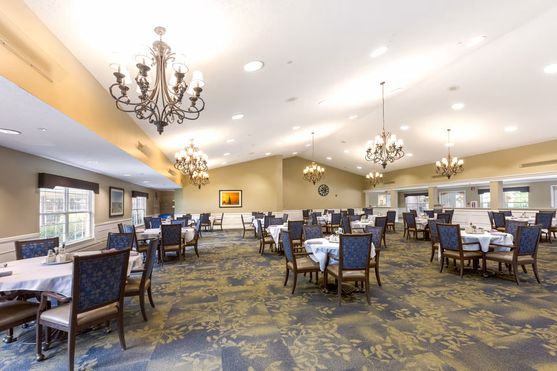 A spacious dining room with multiple round tables covered with white tablecloths, each surrounded by wooden chairs with blue patterned cushions. The room features several ornate chandeliers hanging from the ceiling, large windows letting in natural light, and a carpet with a floral pattern. The walls are painted in a warm beige tone and decorated with framed artwork and a clock.