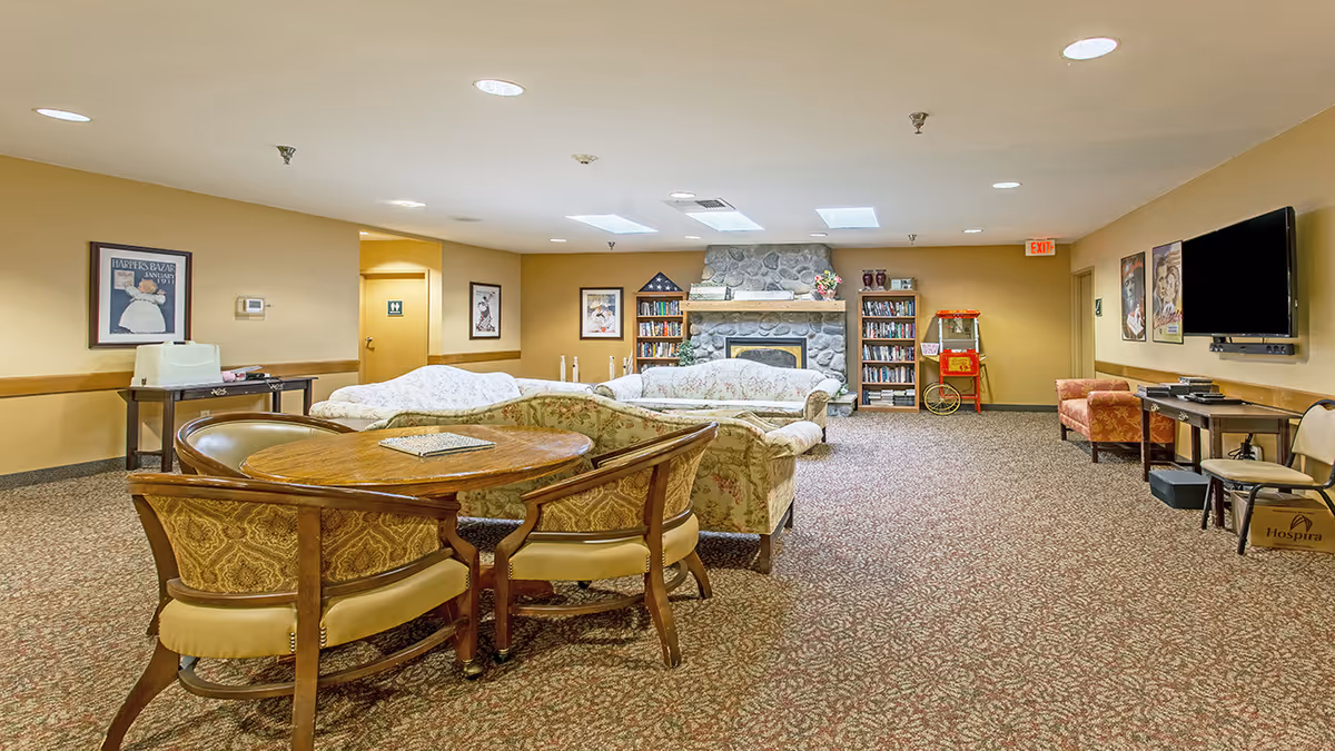 A spacious senior living facility common area with patterned carpet, floral upholstered sofas arranged around a stone fireplace, a round wooden table with four chairs, bookshelves filled with books, a popcorn machine, a wall-mounted TV, and framed artwork on the walls.