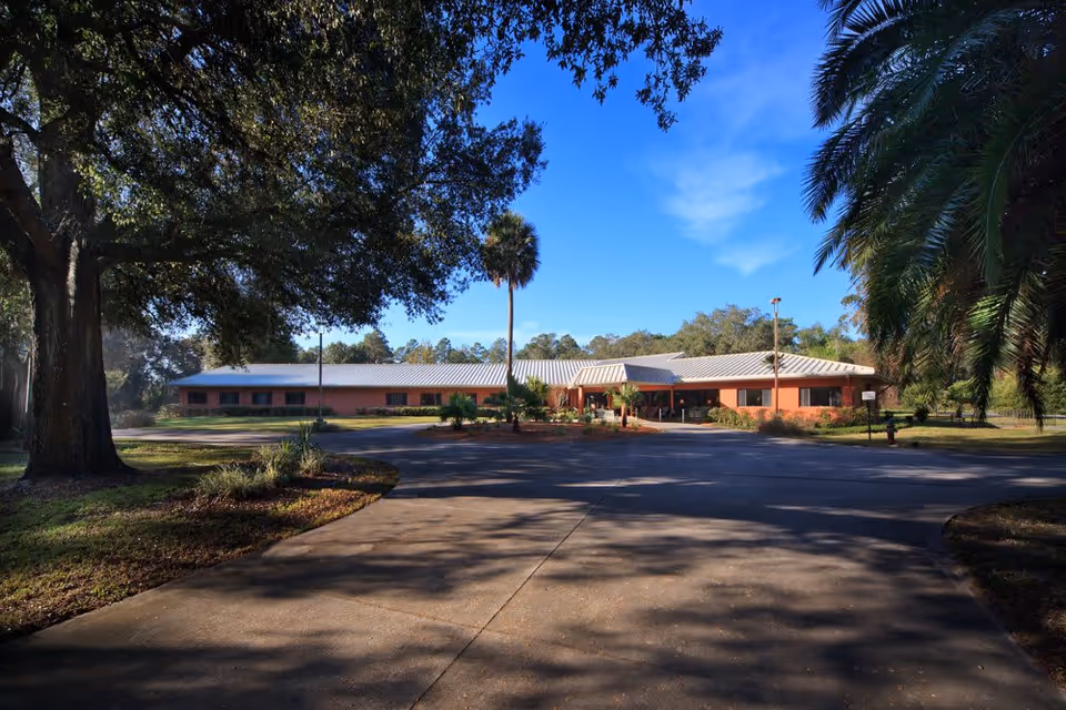 Exterior view of a single-story building with a metal roof surrounded by trees and greenery under a clear blue sky. A paved driveway leads up to the entrance of the building.