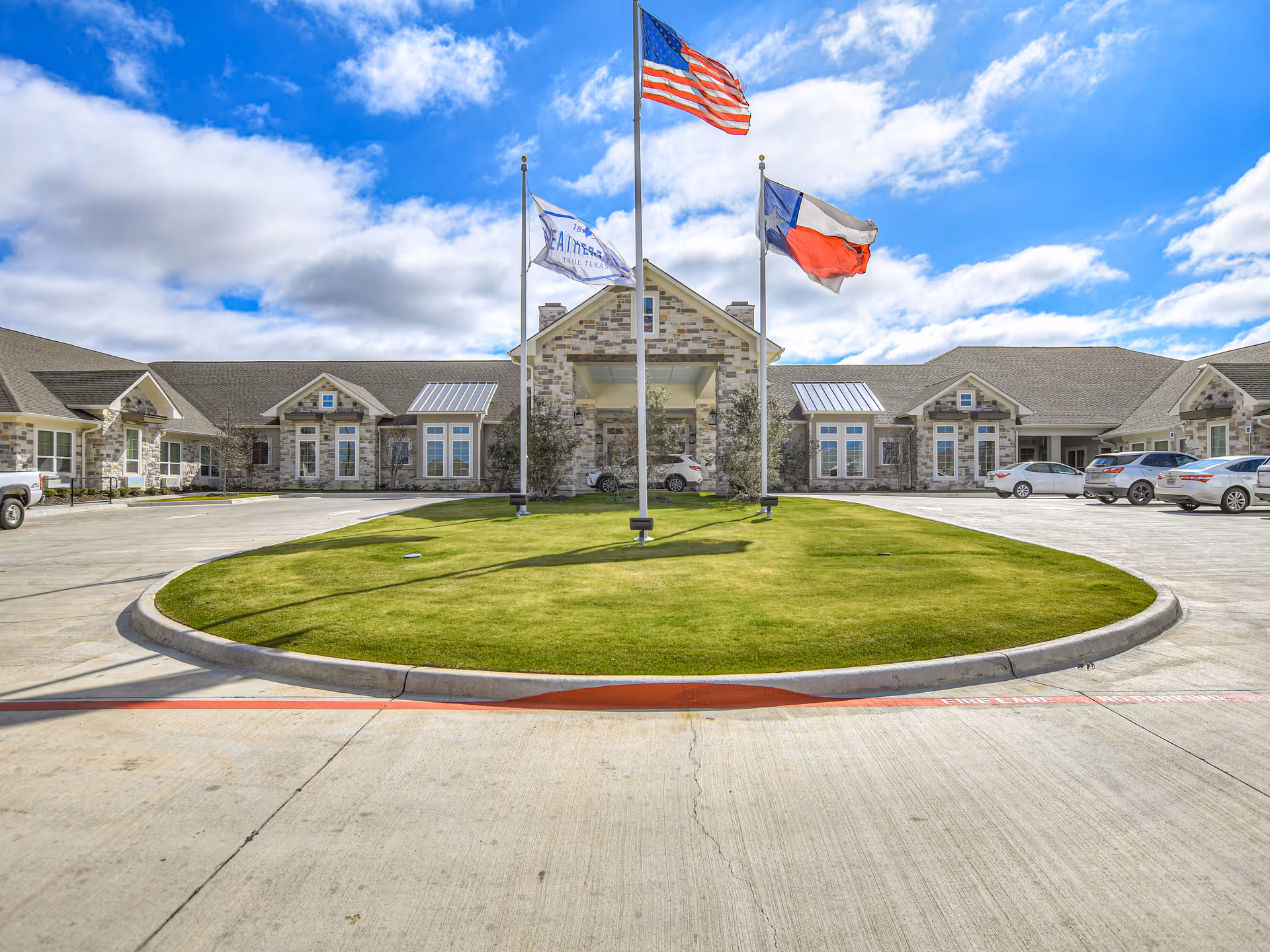 Front exterior view of West Fork at Weatherford facility with a circular driveway, green lawn, and three flagpoles displaying the American flag, Texas state flag, and another flag. Several cars are parked on either side of the driveway under a partly cloudy sky.
