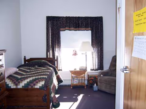 Small furnished bedroom with quilted bed, nightstand, lamp, window, chair, and open door.