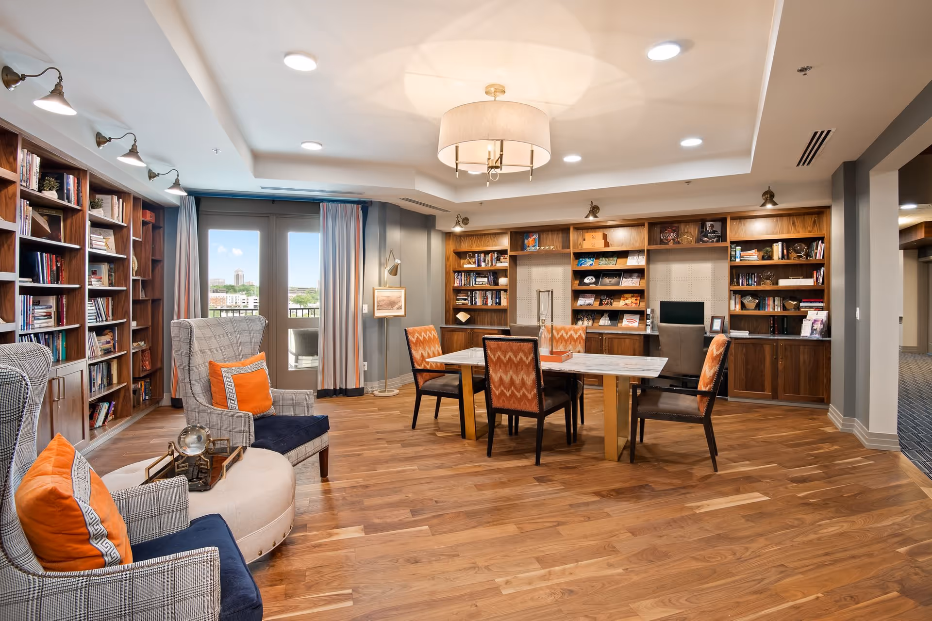 A spacious, well-lit common area in a senior living facility featuring wooden bookshelves filled with books along the walls, a large table with six chairs in the center, and comfortable armchairs with orange cushions. The room has wooden flooring, a modern ceiling light fixture, and large windows with curtains allowing natural light to enter.