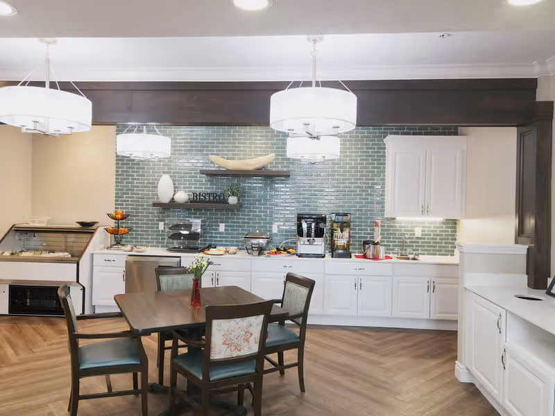 A bright and clean dining area with a wooden table and four chairs, set against a kitchen counter with white cabinets and a green tiled backsplash. The counter has various food and beverage items, including a coffee machine and a juice dispenser. Modern pendant lights hang from the ceiling, illuminating the space.