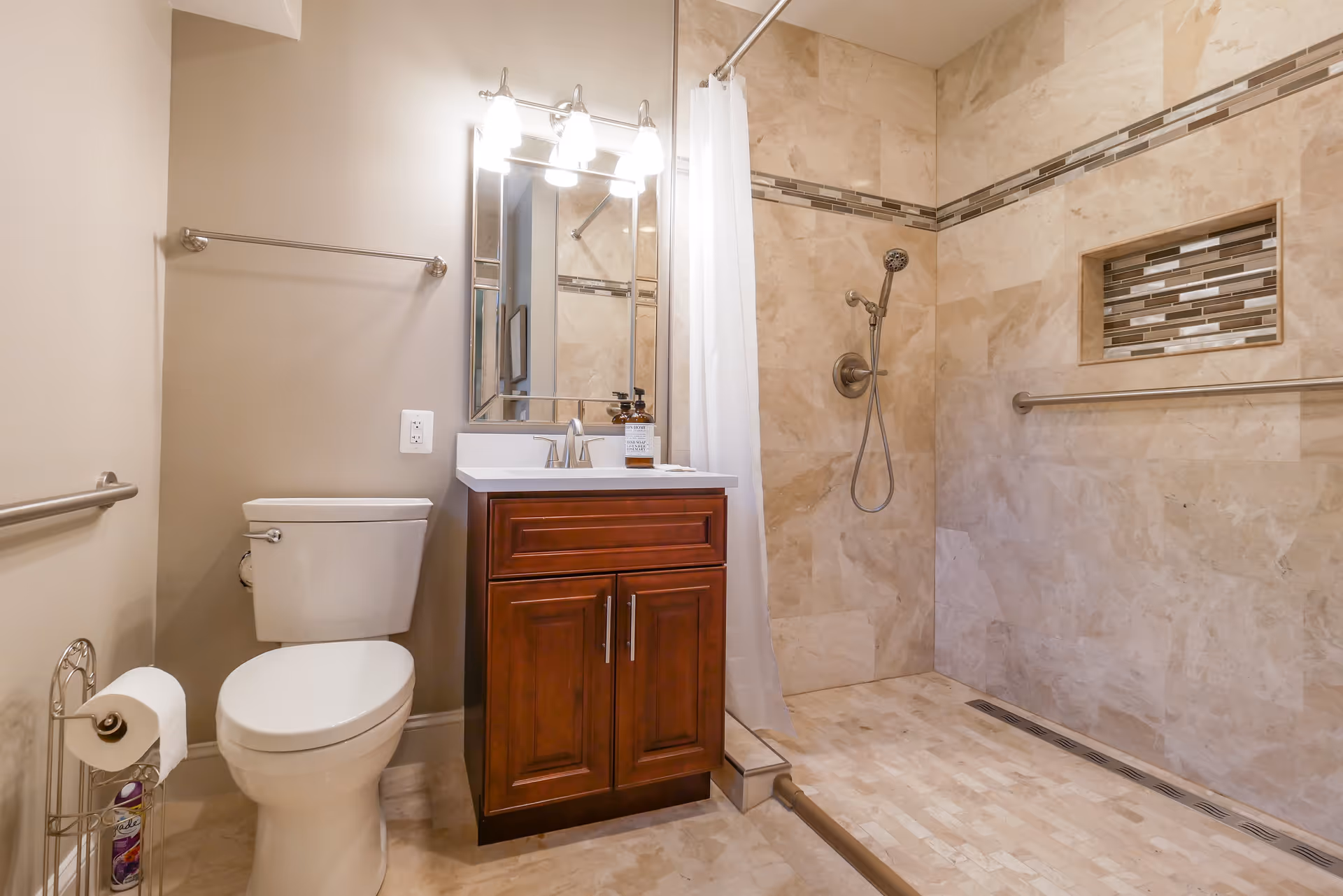 A clean and modern bathroom featuring a white toilet, a wooden vanity with a white countertop and a mirror above it, and a walk-in shower with beige tiled walls and floor. The shower has a handheld showerhead, a built-in niche with decorative tiles, and a white shower curtain. There are metal grab bars on the walls for support and a towel rack above the toilet.