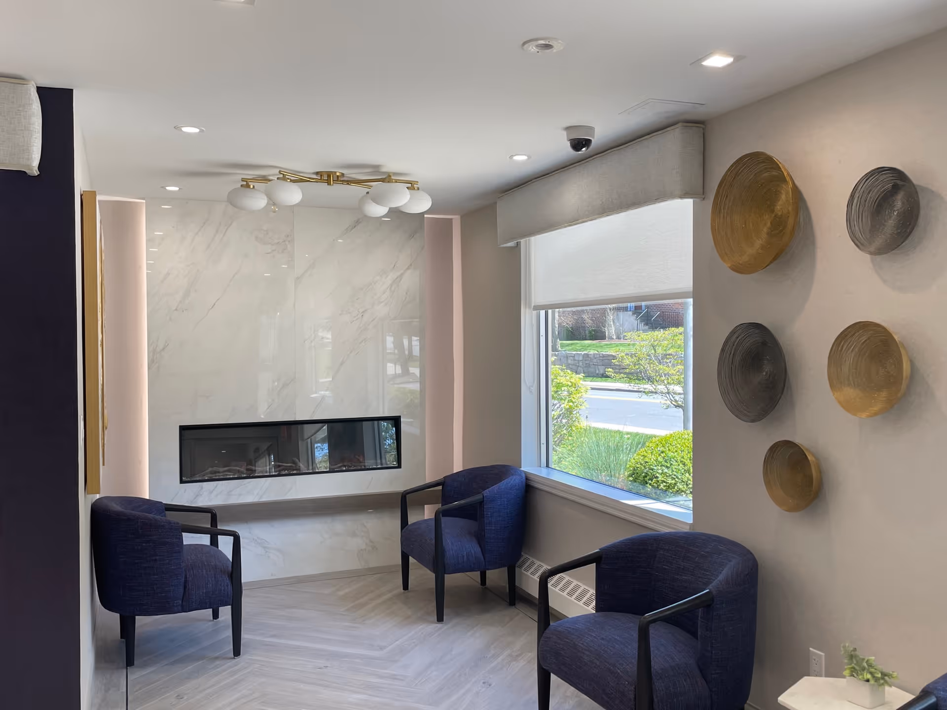 A modern sitting area with three dark blue armchairs arranged around a sleek, built-in electric fireplace set in a white marble wall. A large window with a white roller shade lets in natural light, and decorative round wall hangings in gold and gray tones adorn the adjacent wall. The ceiling has recessed lighting and a contemporary light fixture.