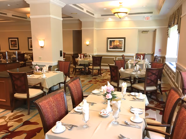 A well-lit dining room with multiple tables covered in beige tablecloths, set with white napkins, cups, plates, and silverware. Each table has a small floral centerpiece. The room features patterned carpet, wall sconces, framed artwork, and large windows with curtains allowing natural light.