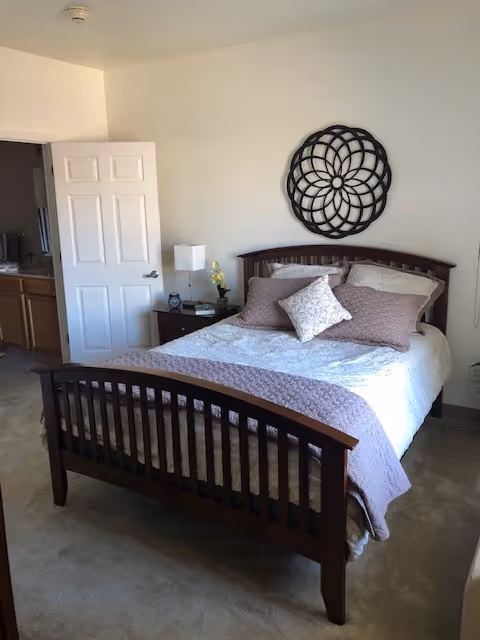 A neatly made bed with a wooden headboard and footboard in a bedroom. The bed has white and lavender bedding with multiple pillows. A nightstand with a lamp, clock, and small plant is next to the bed. A decorative circular wall art piece hangs above the headboard. An open door reveals a glimpse of a bathroom with a sink and mirror.