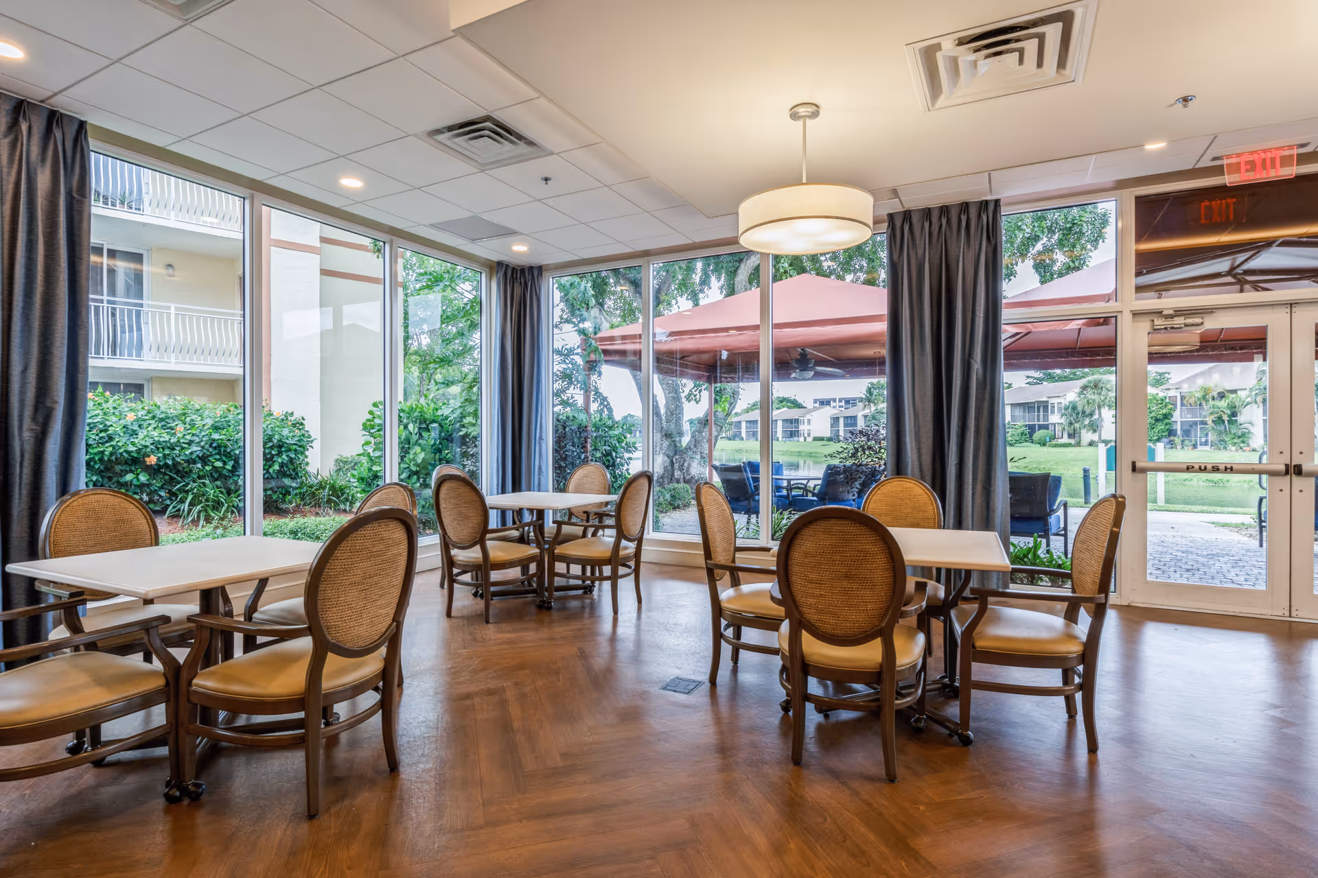 A bright dining room with several tables and chairs arranged neatly. Large floor-to-ceiling windows provide a view of an outdoor patio area with umbrellas and greenery. The room has wooden flooring, gray curtains, and a modern ceiling light fixture.