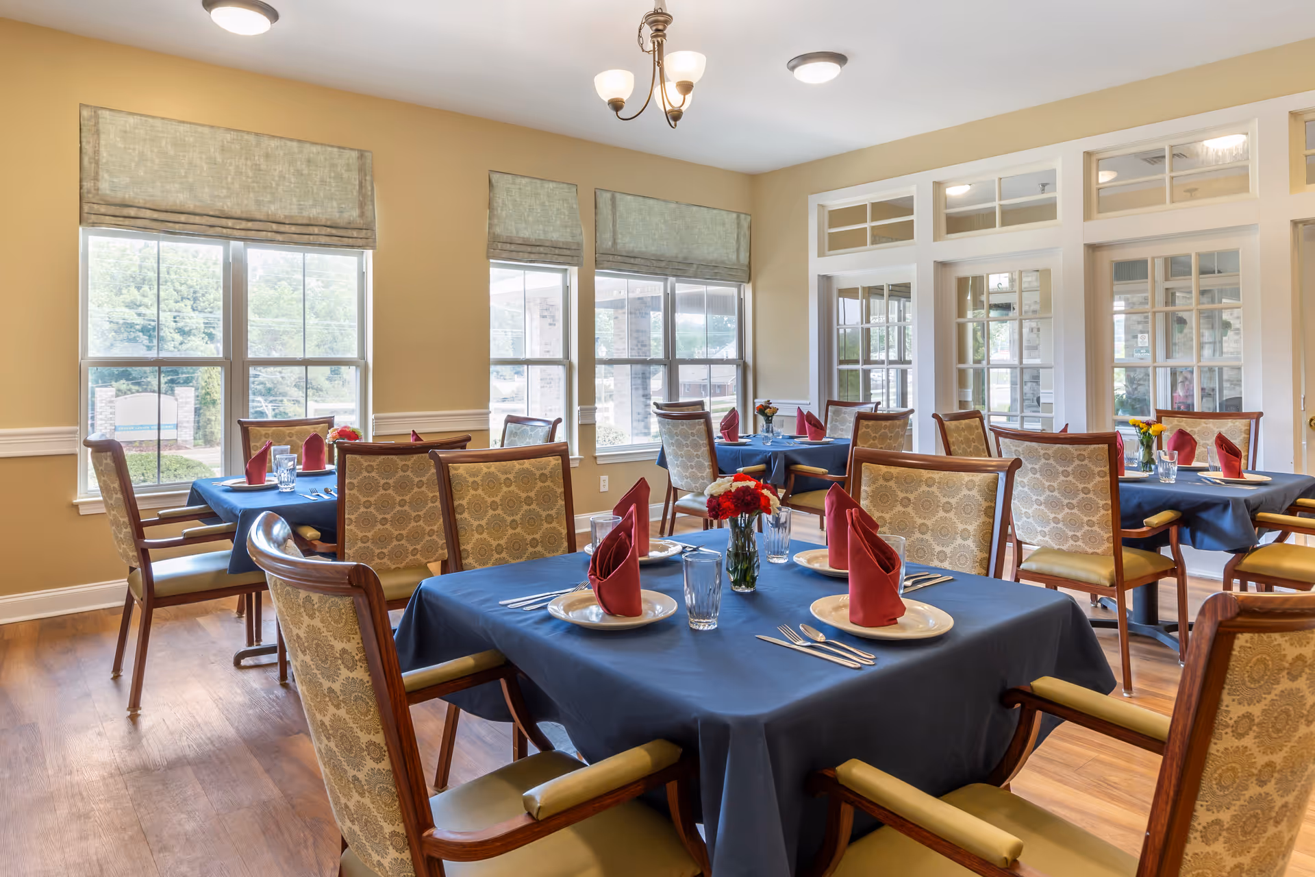 Bright dining room with multiple tables set with blue tablecloths, red napkins, and chairs near large windows.