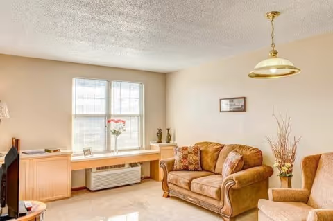 A cozy living room with beige walls and carpet, featuring a brown upholstered loveseat with patterned cushions, a matching armchair, a wooden desk under a window with blinds, decorative vases, a framed picture on the wall, and a hanging ceiling light fixture.