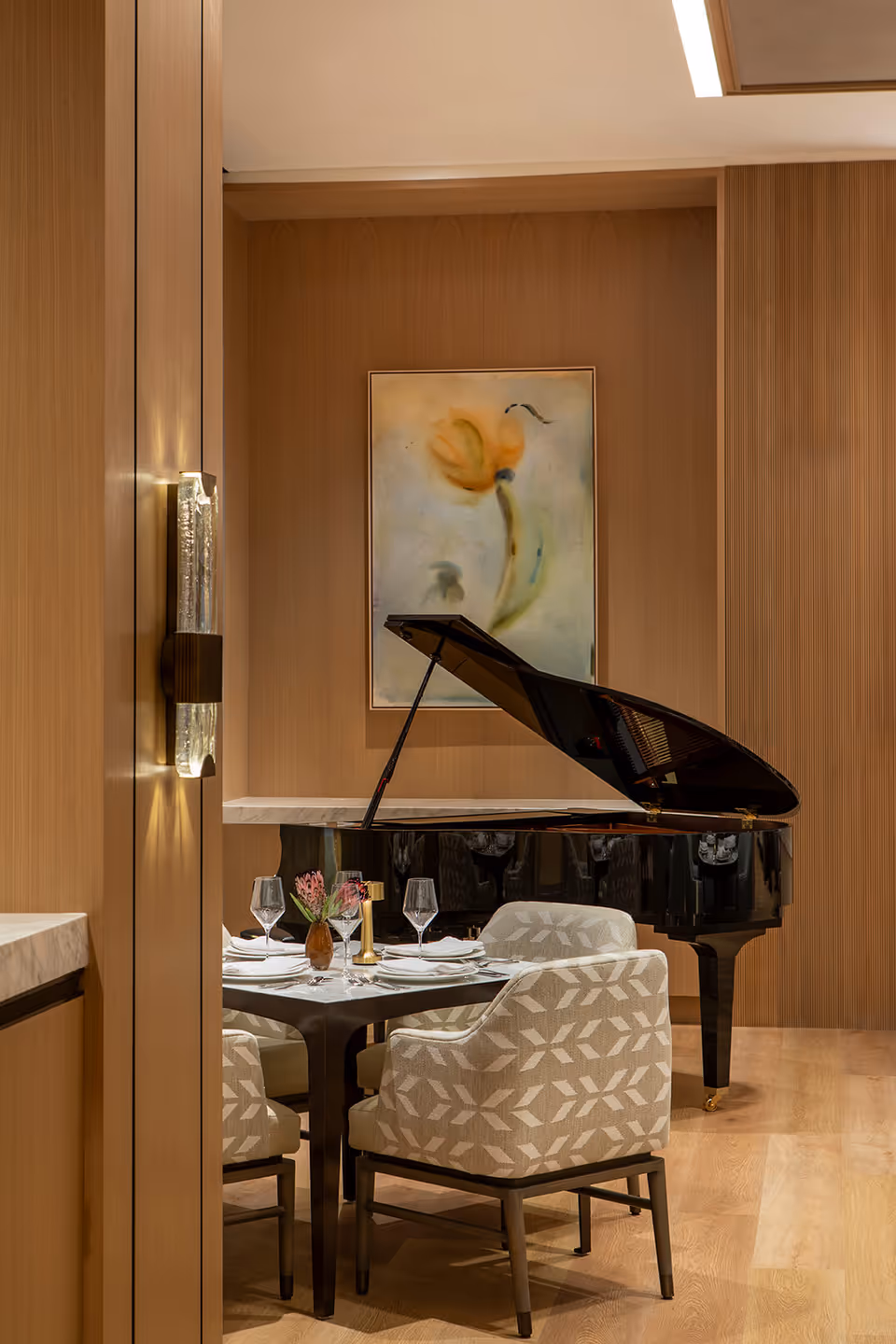 A formal dining area with a set table and upholstered chairs in front of a black grand piano against wood-paneled walls and a floral painting.