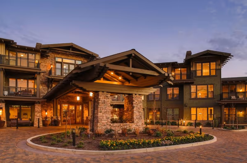 Exterior view of Fountaingrove Lodge (LGBTQ+) at dusk, showing a large, multi-story building with warm interior lights visible through numerous windows. The entrance features a covered driveway with stone pillars and a landscaped roundabout with flowers and shrubs.