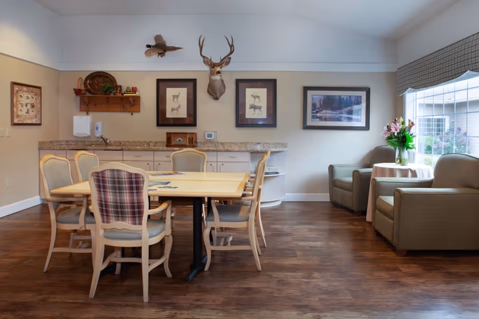 A cozy common area with a wooden table surrounded by six chairs, a countertop with cabinets and a sink in the background, and two armchairs with a small round table holding a vase of flowers near a large window. The walls are decorated with framed pictures and a mounted deer head, and a bird decoration is hanging from the ceiling.