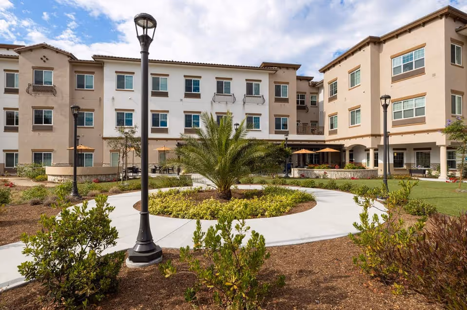 Exterior courtyard of a three-story beige senior living building with a circular walkway, landscaping, palm tree, lampposts, and patio umbrellas.