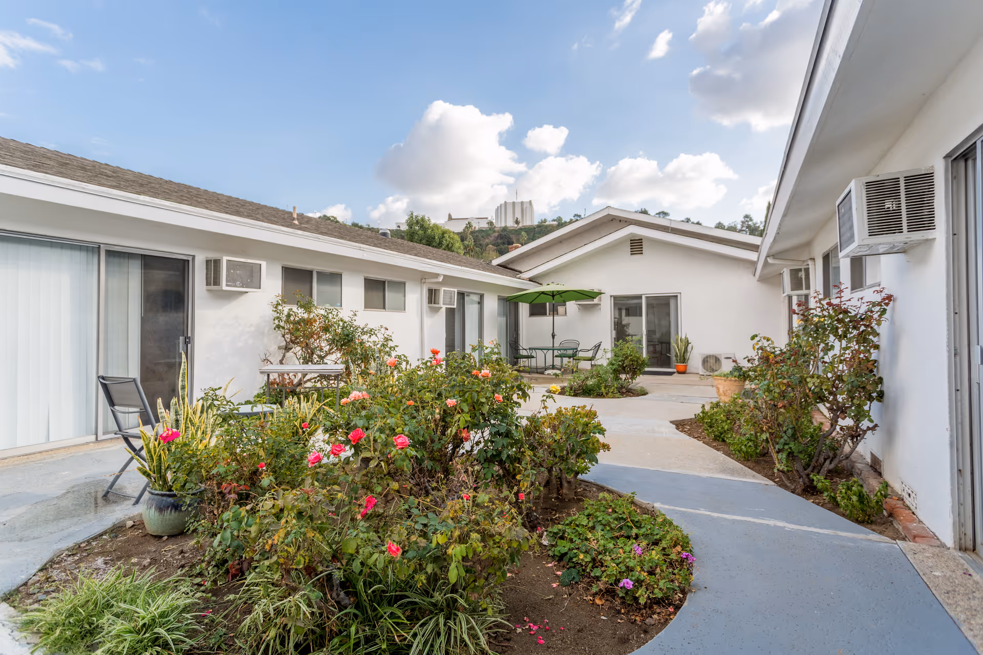 Outdoor courtyard area of a senior living facility with white buildings surrounding a garden filled with various flowering plants and shrubs. There are sliding glass doors, window air conditioning units, a green patio umbrella with chairs and tables, and a clear blue sky with some clouds above.