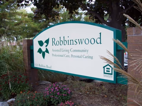Outdoor sign for Robbinswood Assisted Living Community surrounded by plants and trees. The sign includes the text 'Robbinswood Assisted Living Community Professional Care, Personal Caring' and the address '1125 Robbins Road'. There is also a logo indicating equal housing opportunity.