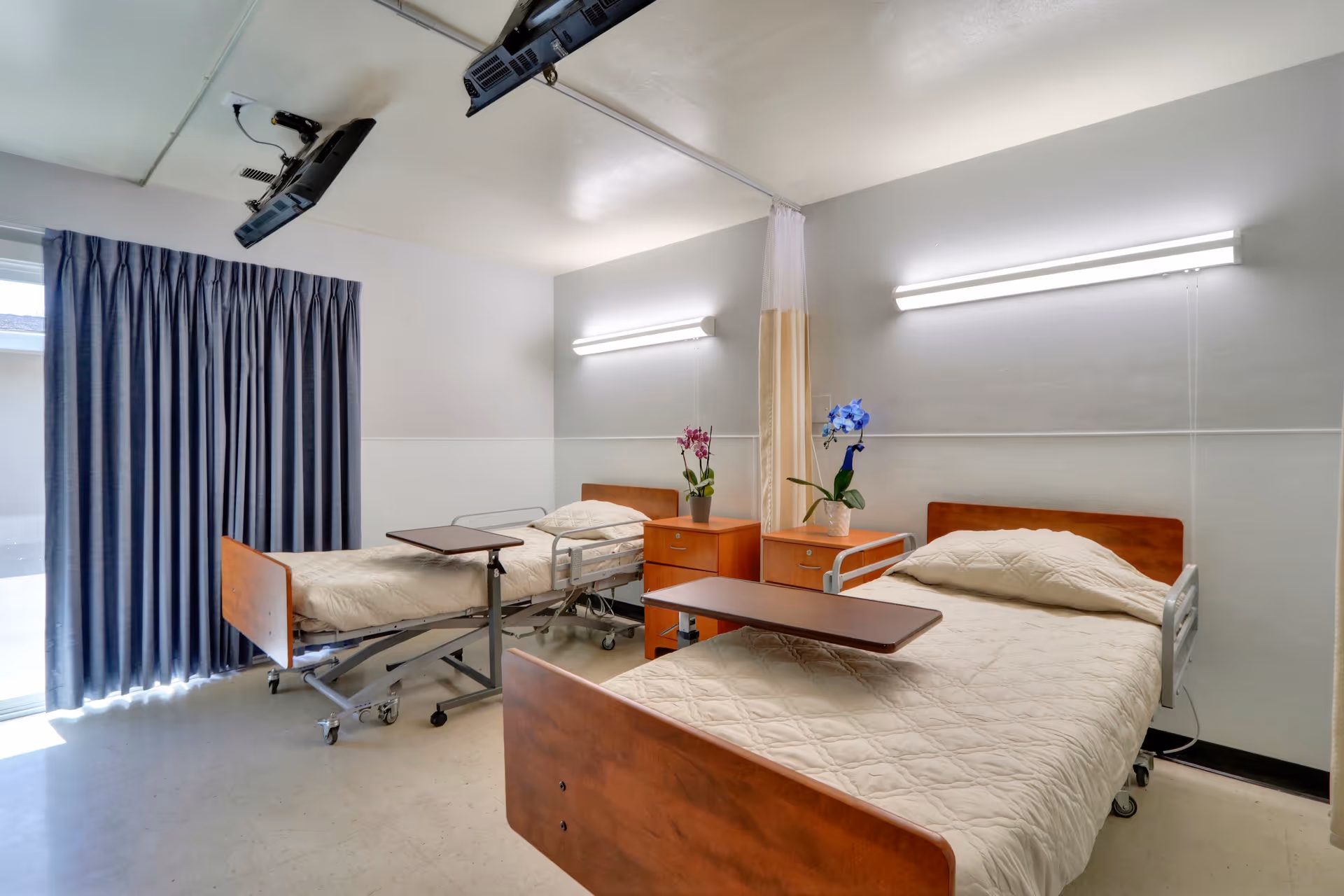 Two hospital-style beds with wooden headboards and overbed tables in a bright shared patient room with nightstands, wall lights, and a privacy curtain.