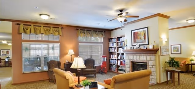 A cozy living room area in a senior living facility with comfortable seating including a sofa and armchairs arranged around a coffee table. There is a stone fireplace with a wooden mantle, a bookshelf filled with books, a ceiling fan with lights, and large windows with valances allowing natural light to enter. The room is warmly decorated with lamps, framed artwork, and plants.