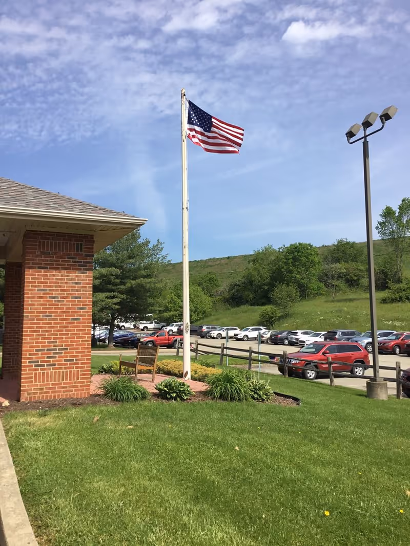 Outdoor scene showing a flagpole with an American flag waving, a brick building corner, a wooden bench surrounded by plants, a parking lot with multiple cars, a tall streetlight, and a grassy hill with trees under a partly cloudy sky.