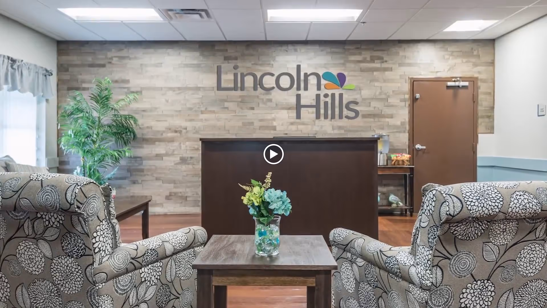 A cozy lounge area in Lincoln Hills facility featuring two patterned armchairs facing a small wooden table with a vase of flowers. Behind the table is a dark wooden counter against a textured stone wall with the Lincoln Hills logo displayed. A green potted plant is on the left side near a window with curtains, and a closed brown door is on the right side.