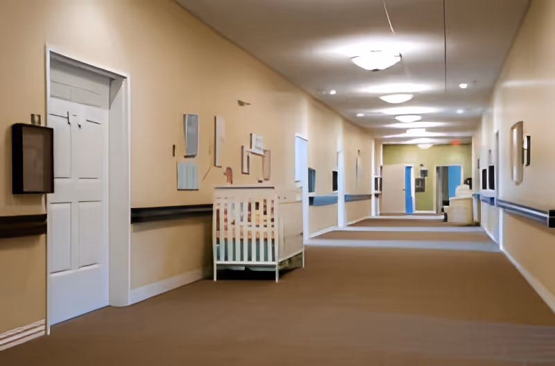 A long, well-lit hallway in a senior living facility with beige walls and carpeted floor. The hallway features handrails on both sides, several closed doors, framed pictures on the walls, and a baby crib placed against one wall. Ceiling lights evenly illuminate the corridor.