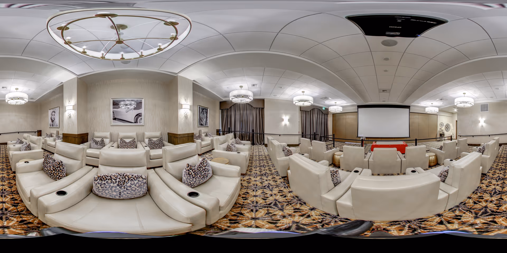 A spacious communal media room with rows of cream leather recliners, patterned carpet, decorative pillows, chandeliers, and a large projection screen at the front.