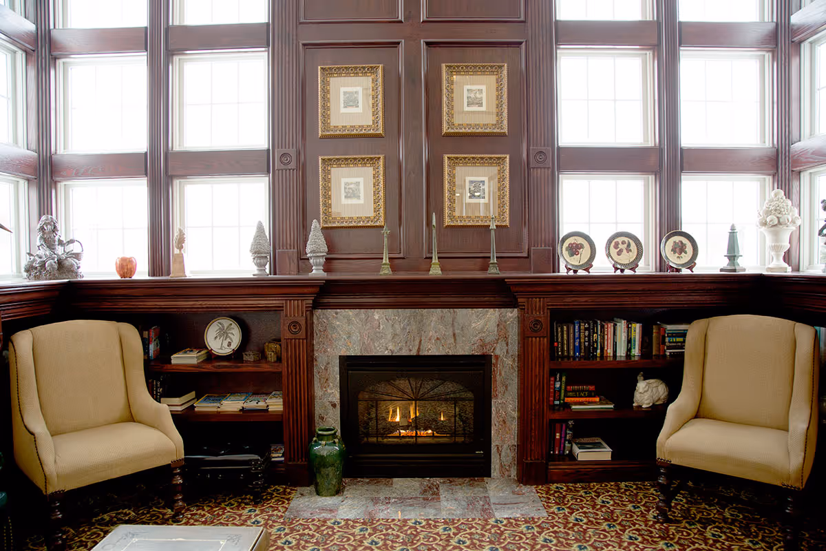 A cozy living room area with two beige armchairs facing a fireplace with a marble surround. The fireplace is set in a dark wood paneled wall with four framed pictures above it. Built-in bookshelves flank the fireplace, holding books and decorative items. Large windows with multiple panes surround the upper part of the wall, letting in natural light. The floor has a patterned carpet.