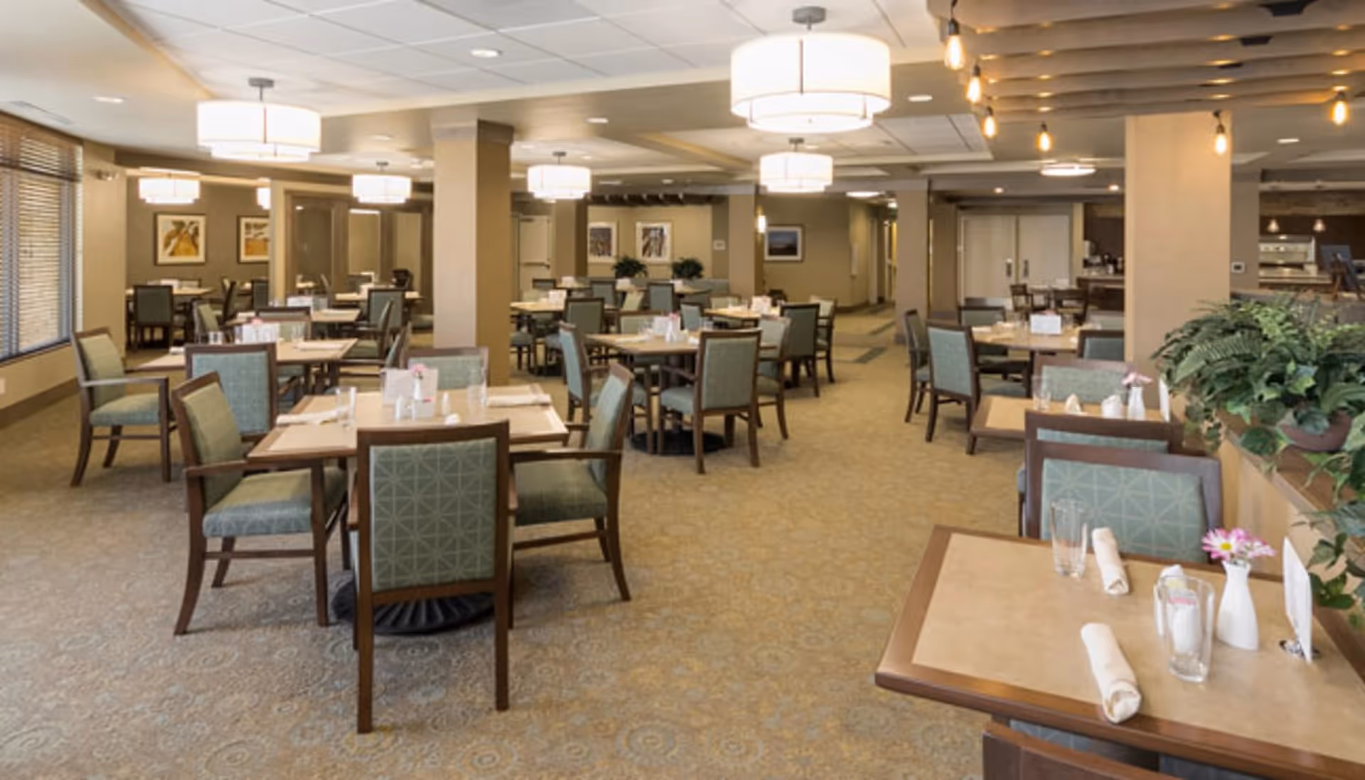A spacious dining room with multiple square wooden tables and green cushioned chairs arranged neatly. Each table is set with rolled napkins, glasses, and small flower vases. The room is well-lit with modern ceiling lights and has large windows on one side allowing natural light. There are plants and framed artwork on the walls, creating a warm and inviting atmosphere.