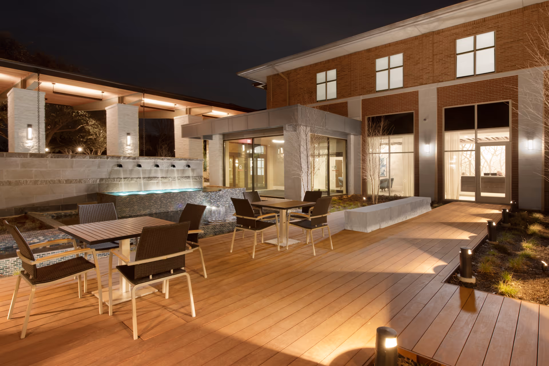 Outdoor patio area at night with wooden decking, tables and chairs, a water feature with multiple spouts, and a building with large windows illuminated from inside.