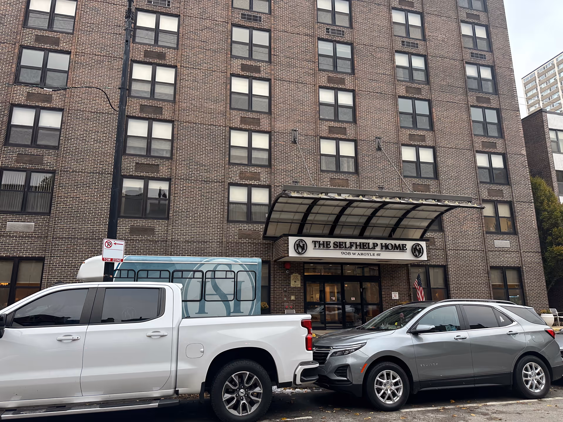 Exterior view of The Selfhelp Home Senior Living Community building showing a multi-story brick facade with many windows. Two vehicles, a white pickup truck and a gray SUV, are parked in front of the entrance. The entrance has a canopy with the facility's name and address displayed above the doors.