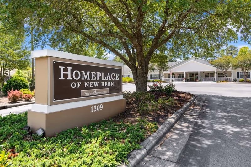 Entrance sign for Home Place of New Bern with landscaping and a large tree, with the facility building visible in the background under a clear blue sky.