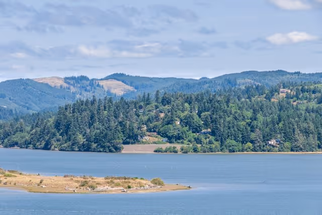View across a blue lake to a tree-covered shoreline and distant forested hills under a partly cloudy sky.