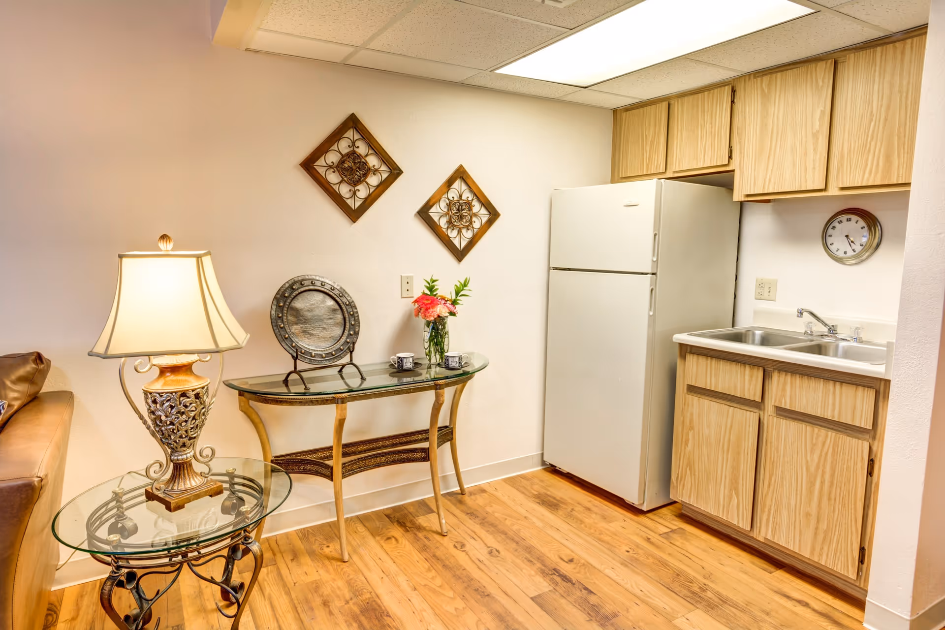 Cozy kitchenette and living nook with a refrigerator, sink and wood cabinets beside glass side tables and a lamp.