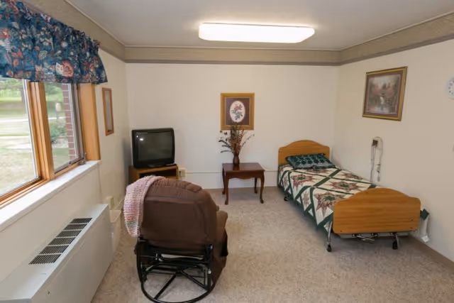 Small resident bedroom with a single bed, recliner, TV on a stand, side table, and a large window.