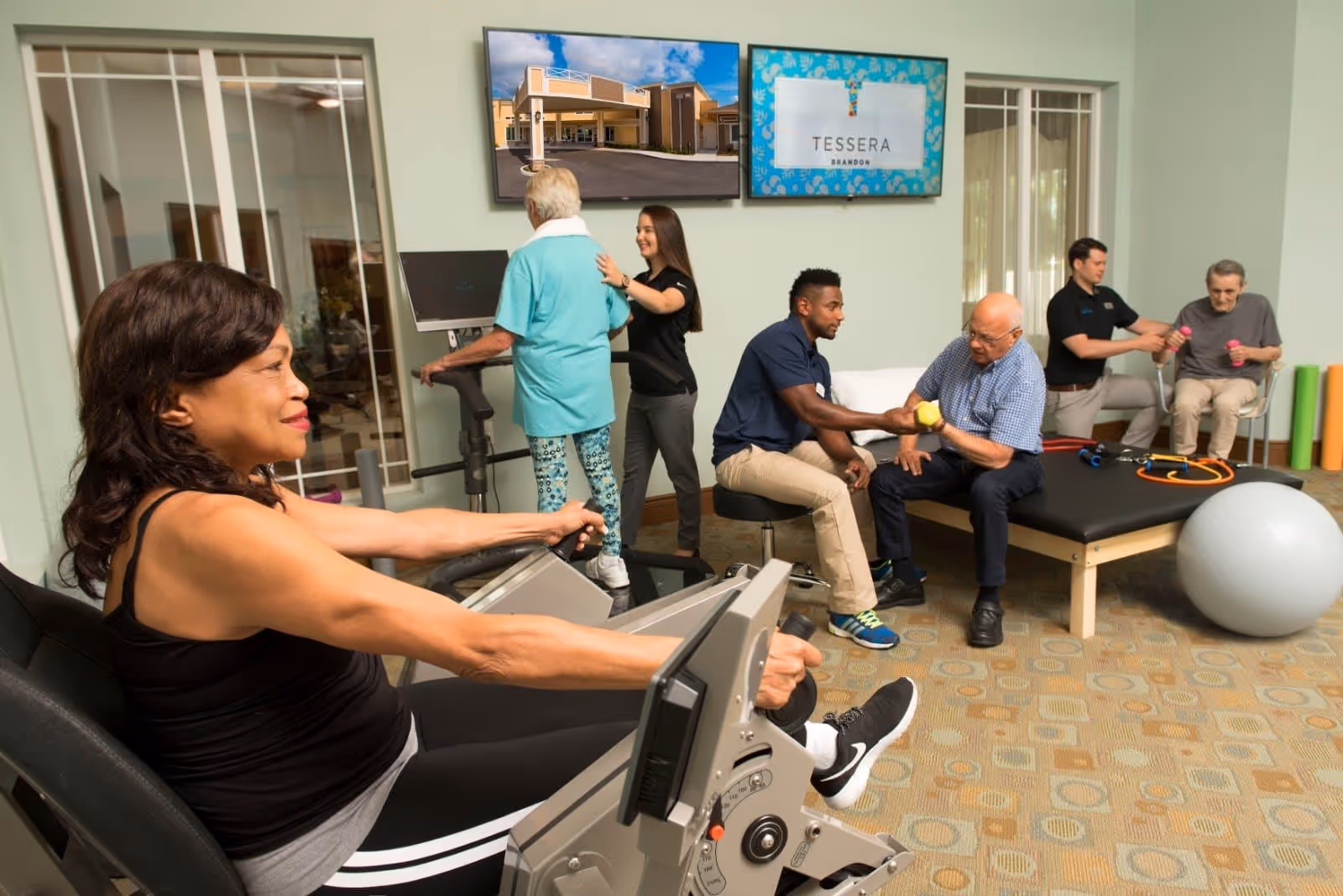 A senior living facility exercise room where elderly individuals are engaging in physical therapy and fitness activities with the assistance of staff. One woman is using a rowing machine, an elderly woman is on a treadmill with a staff member assisting, a man is lifting a yellow dumbbell with a trainer, and another elderly man is using pink dumbbells with help from another staff member. Two screens on the wall display an exterior image of the facility and the Tessera of Brandon logo.